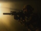 A soldier in tactical gear aims a weapon with a laser sight in low light conditions.