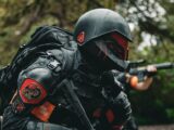 A soldier in advanced tactical armor with a firearm crouches in a forested area during a mission.