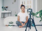 Smiling woman filming a vlog in cozy indoor setting with plants and soft lighting.