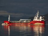 A vibrant red cargo ship sails on the ocean at twilight with mountains in the background.