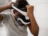 A man in a gray shirt adjusting his virtual reality headset inside a bright room.