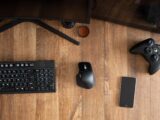 Top view of black gamepad with mouse and keyboard on wooden desk with monitor in apartment in daytime