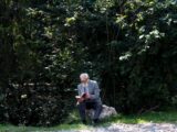 Senior man with gray hair reading a book outside in a tranquil forest environment.