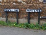 Two street signs, Manor Lane and Park Lane, on a rustic stone wall backdrop.