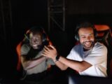 Two young male gamers celebrating a victory with a handshake and smiles in a dark gaming room.